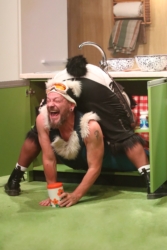 A stage scene from The Three Bears. Panda bear is bending over, looking inside a kitchen cupboard, all we can see are his legs and furry tail. Polar bear is popping out of the same kitchen cupboard through Panda's legs, all we can see are his arms and head. Polar bear is laughing and holding a jar of porridge oats.