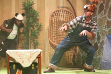 A stage scene from The Three Bears. Grizzly bear, wearing dungarees and a miner's hat with brown fur trim, looks warily at a strange box under a small wooden table. In the background hiding behind a tall plant is Panda bear wearing black and white clothes with a black fur trim.