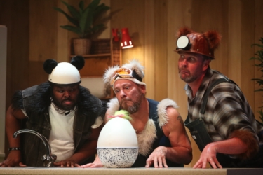 A stage scene from The Three Bears. Three performers are behind a kitchen cabinet. Panda, a bear inspired by a panda, is wearing black and white clothes with a black fur trim. Polar, a bear inspired by a polar bear, is wearing a blue ski suit, ski hat and goggles with white fur trim. Grizzly, a bear inspired by a grizzly bear, wears dungarees and a miner's hat with brown fur trim. They are all looking bemused at a large egg with a yellow tuft of fluff and a red gingham bow on the top which is in front of them.