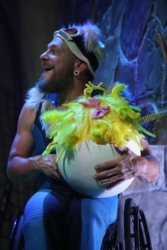 A stage scene from The Three Bears. Polar, a bear inspired by a polar bear, is wearing a blue ski suit, ski hat and goggles with white fur trim. He has an excited look on his face and is sat in his wheelchair holding a large round white half eggshell, inside the egg is ‘Goldie’ a mass of bright yellow feathers with a red bow. Polar is sat infront of a stone-textured backdrop with dramatic stage lighting.